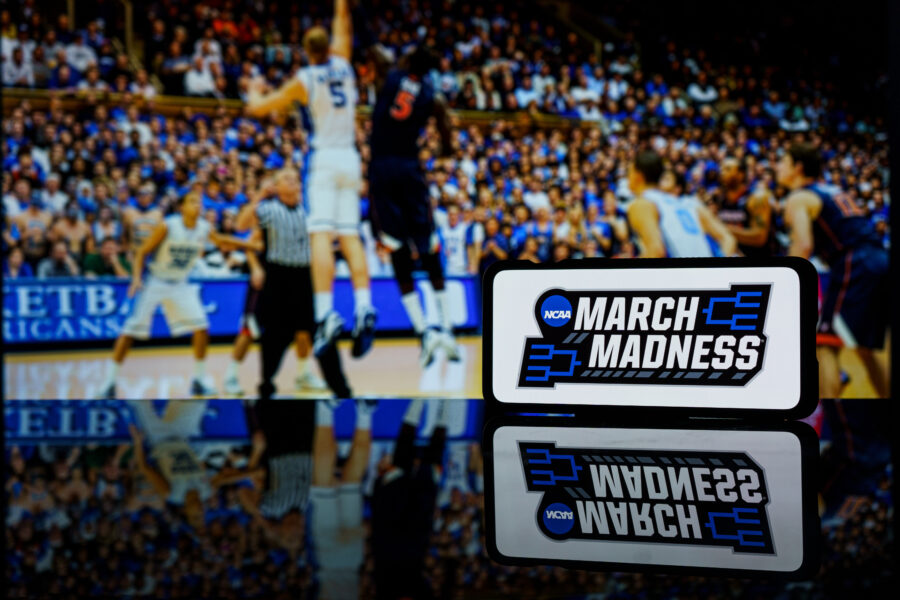 March Madness logo displayed on a smartphone with a live NCAA basketball game in the background.